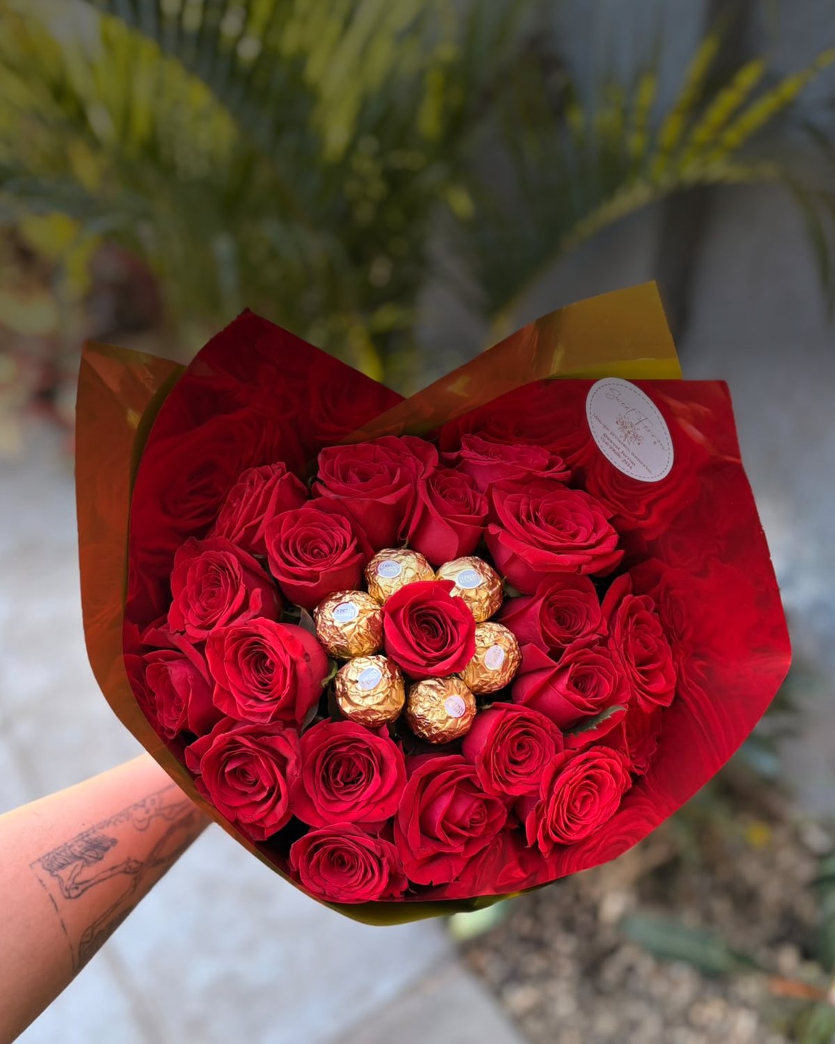 Buquê de rosas vermelhas da Sweet Ferron com bombons Ferrero Rocher no centro, embrulhado em papel vermelho, segurado pela florista, com o jardim ao fundo.