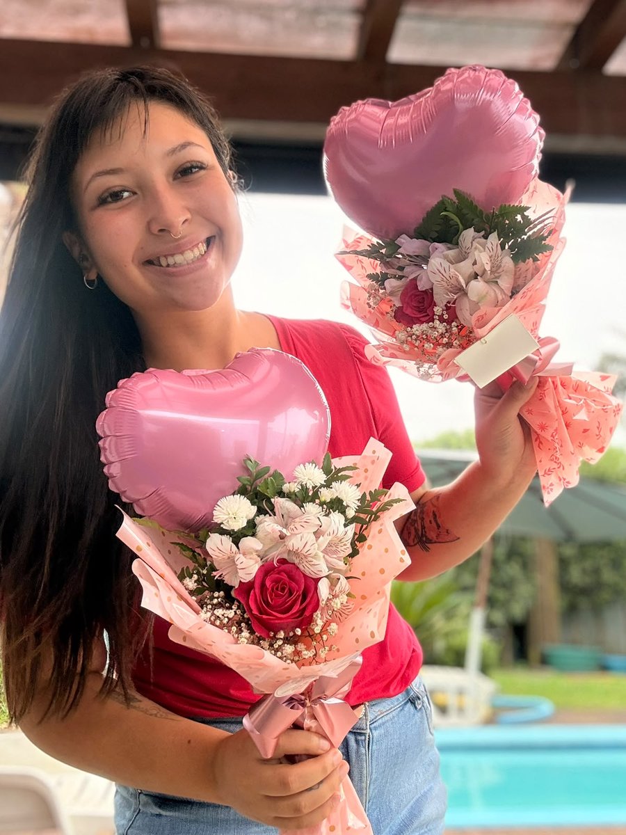 Hevelin sorrindo, segurando dois buquês de flores com balões de coração.
