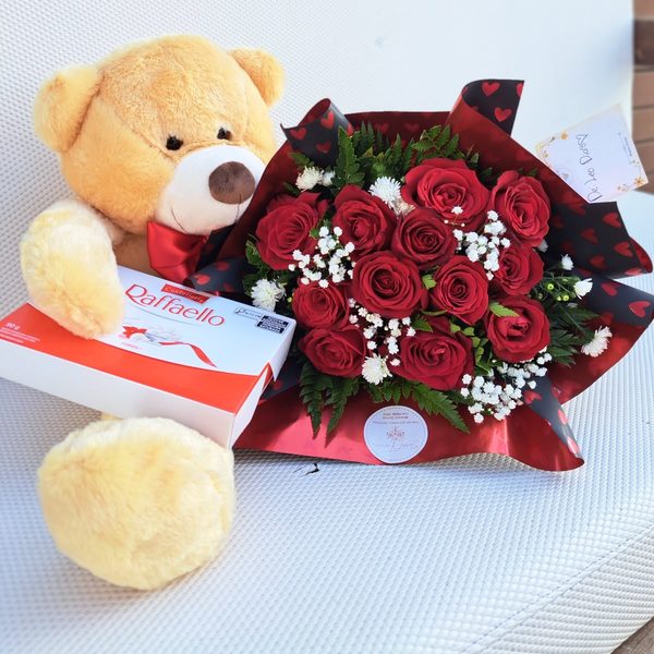 Buquê de rosas vermelhas com mosquitinho ao lado de urso de pelúcia bege e caixa de bombons Raffaello.