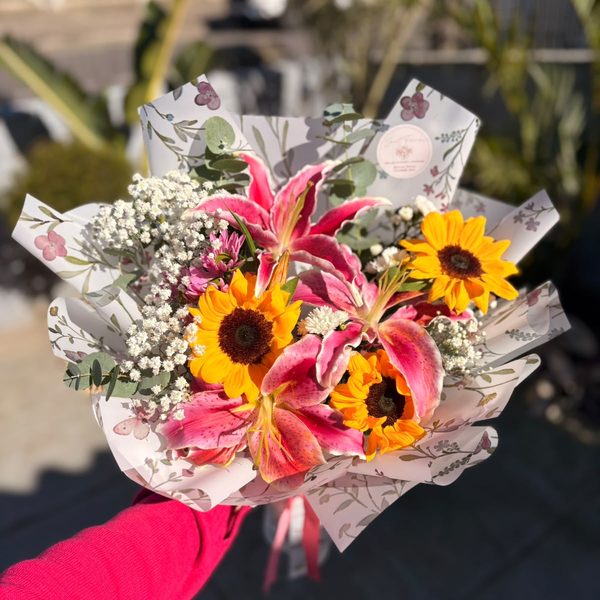 Buquê artesanal de girassóis e lírios rosa com mosquitinho, embrulhado em papel floral, segurado pela florista.