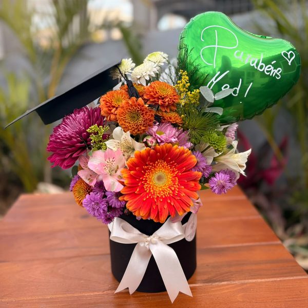 Arranjo de formatura em caixa preta com capelo, gérbera laranja, crisântemos coloridos e balão coração verde escrito Parabéns.